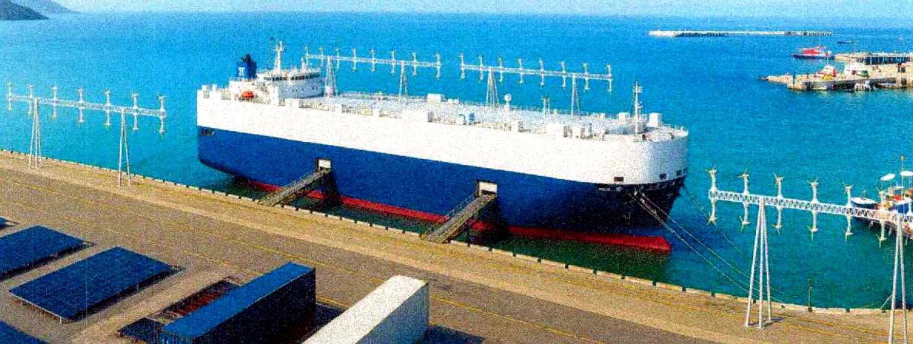 A vessel moored at a quay carrying rows of FlowGen turbine frames, with solar panels and containers on the dockside.